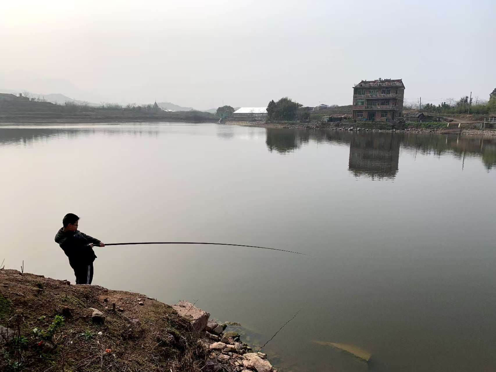 A child fishing from a dirt bank on a still lake