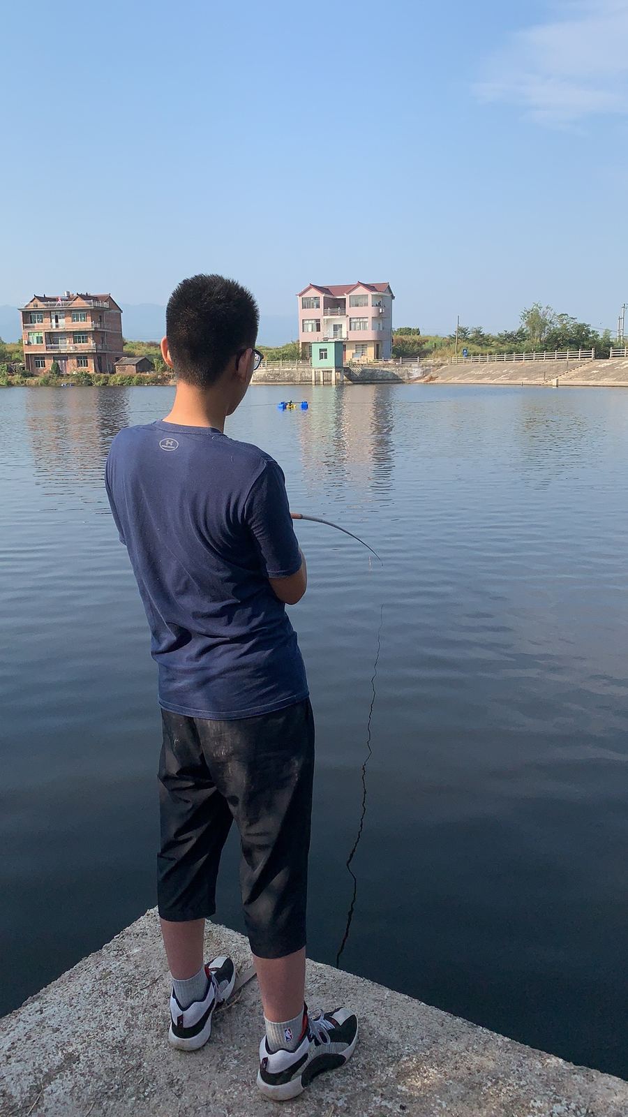 Tianqiqi fishing from a concrete edge, facing houses across the water