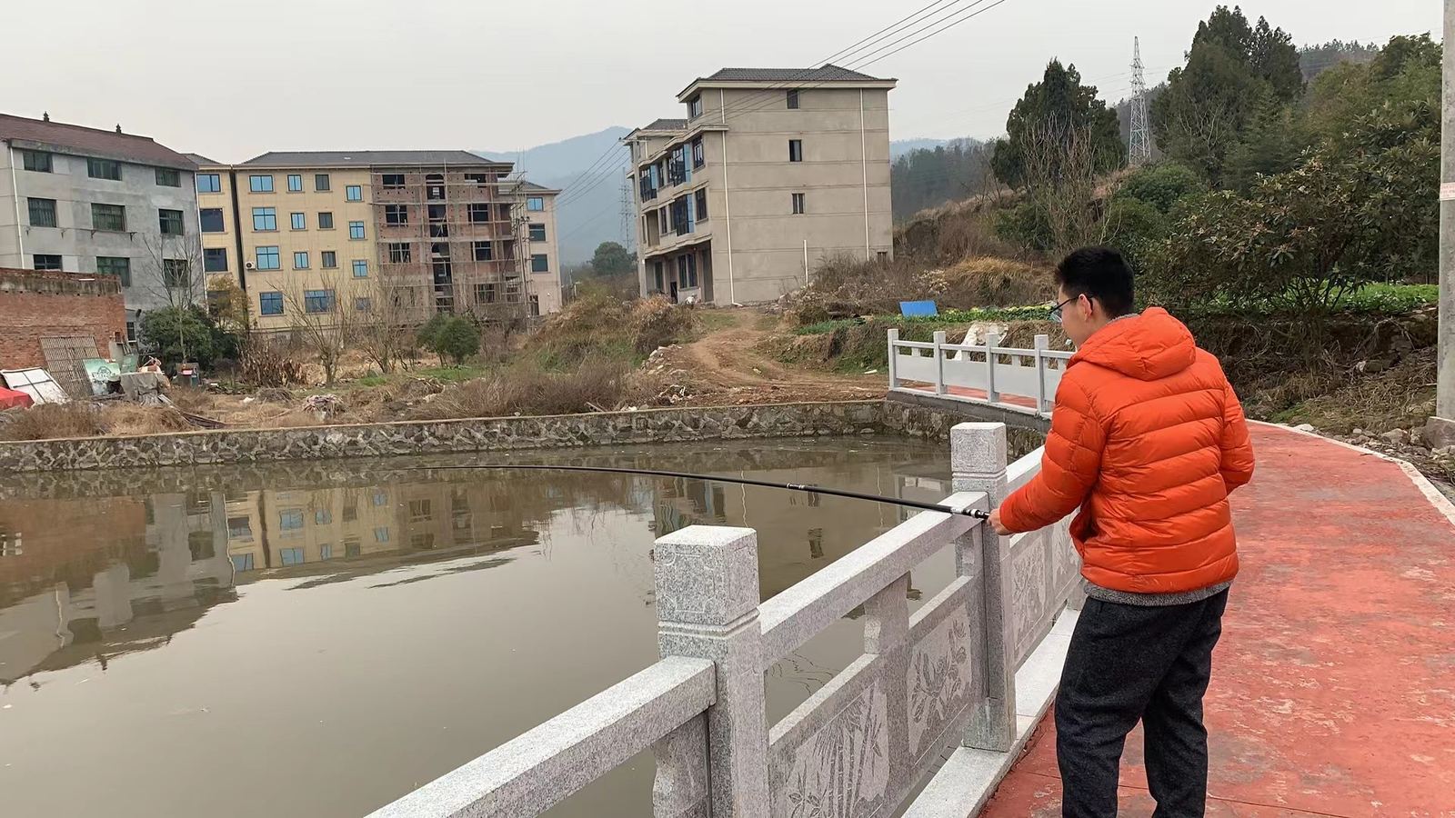 Tianqiqi in an orange jacket fishing from a stone bridge